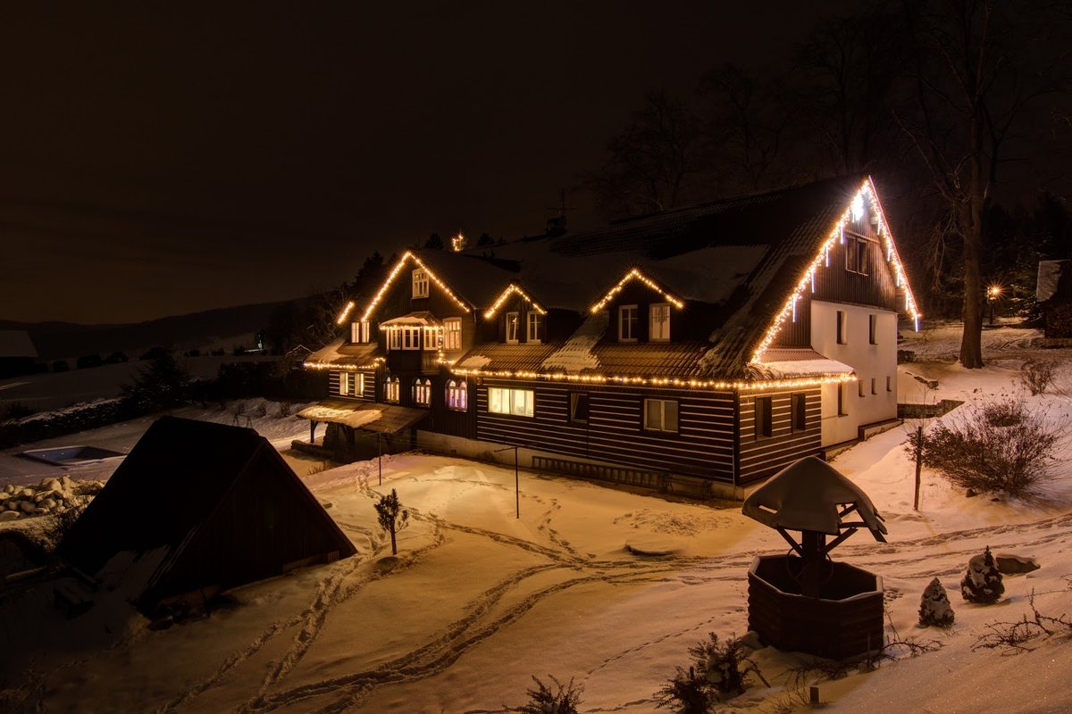 Holzhaus im Schnee bei Nacht mit warmer LED-Lichterkette an der Dachkante