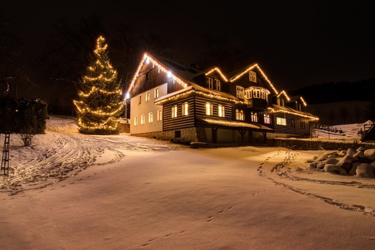Holzhaus bei Nacht mit Lichterkette an der Dachkante und beleuchtetem Tannenbaum im Schnee